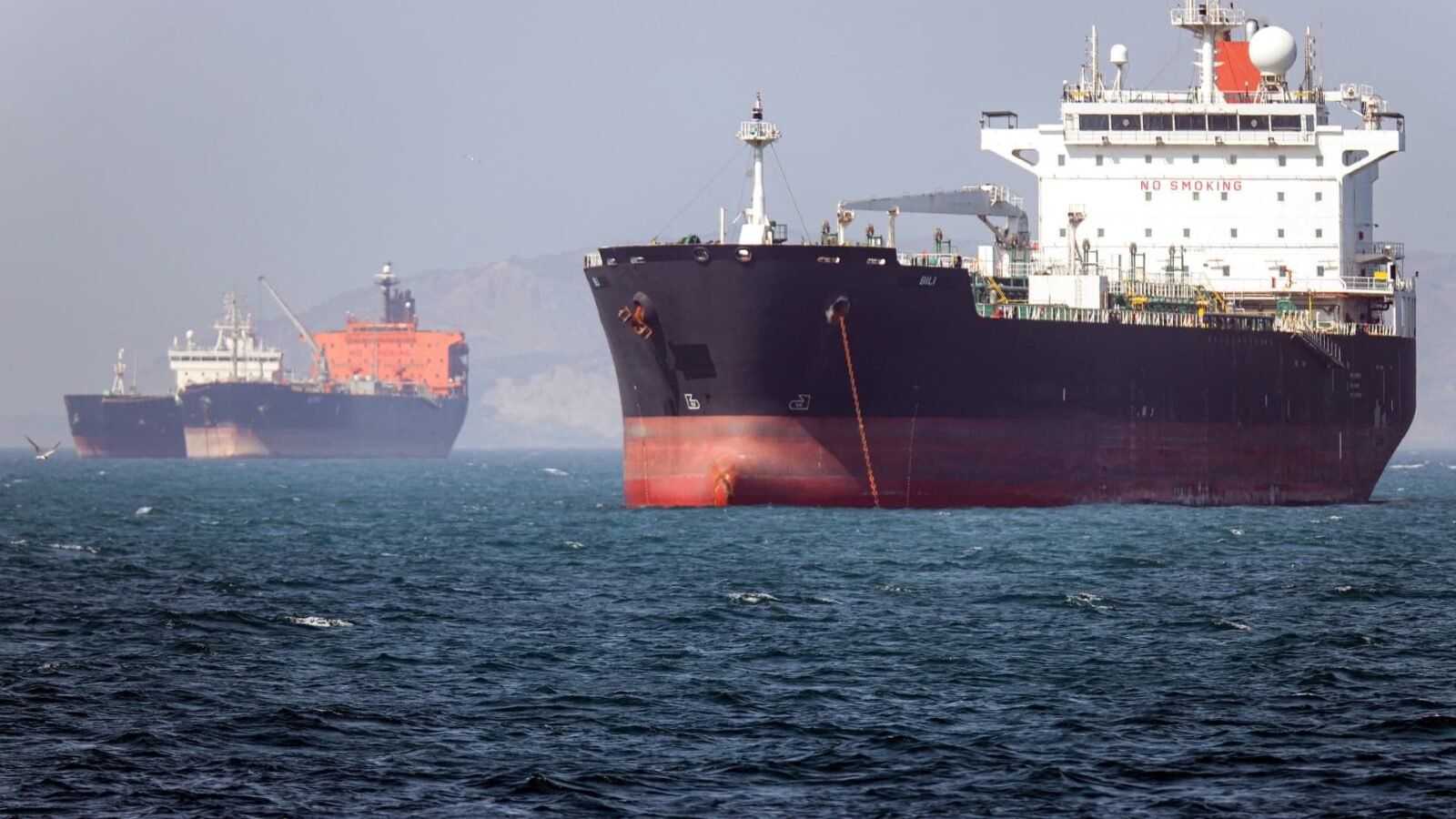 Oil tanker navigating the Strait of Hormuz with a naval vessel in the background, symbolizing the precarious security situation.
