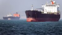 Oil tanker navigating the Strait of Hormuz with a naval vessel in the background, symbolizing the precarious security situation.