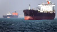 Oil tanker navigating the Strait of Hormuz with a naval vessel in the background, symbolizing the precarious security situation.